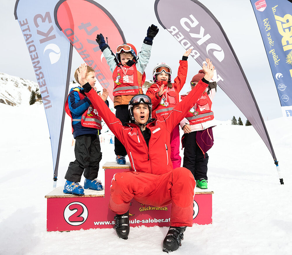 Vier kinderen staan op het podium na de skirace en juichen, een skileraar zit voor hen met uitgestrekte armen en juicht ook