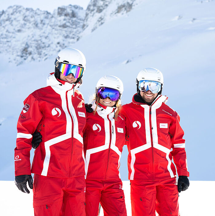 Three ski instructors with reflective goggles are standing arm in arm on the slopes